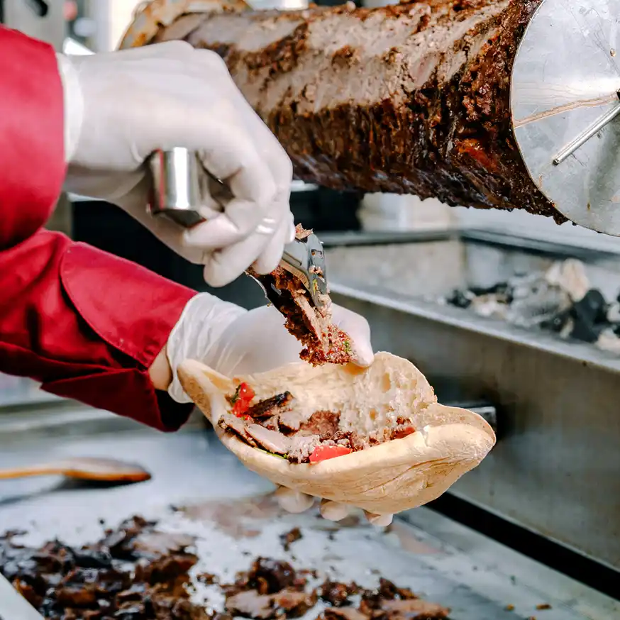 Człowiek w bordowej marynarce trzyma w rękach zawiniętego w papier kebaba. Scena wygląda na przytulną i swobodną, przekazując uczucie ciepła i komfortu.