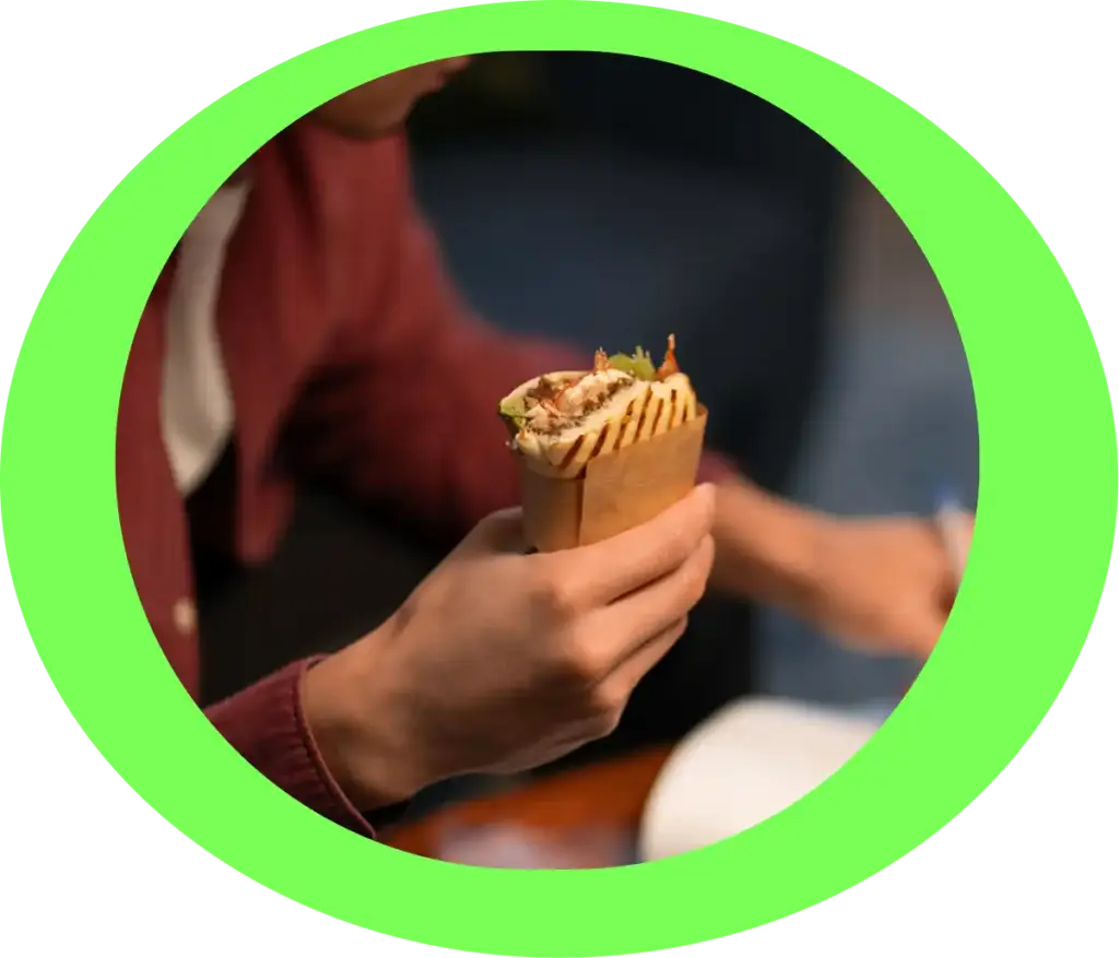 Człowiek w bordowej marynarce trzyma w rękach zawiniętego w papier kebaba. Scena wygląda na przytulną i swobodną, przekazując uczucie ciepła i komfortu.