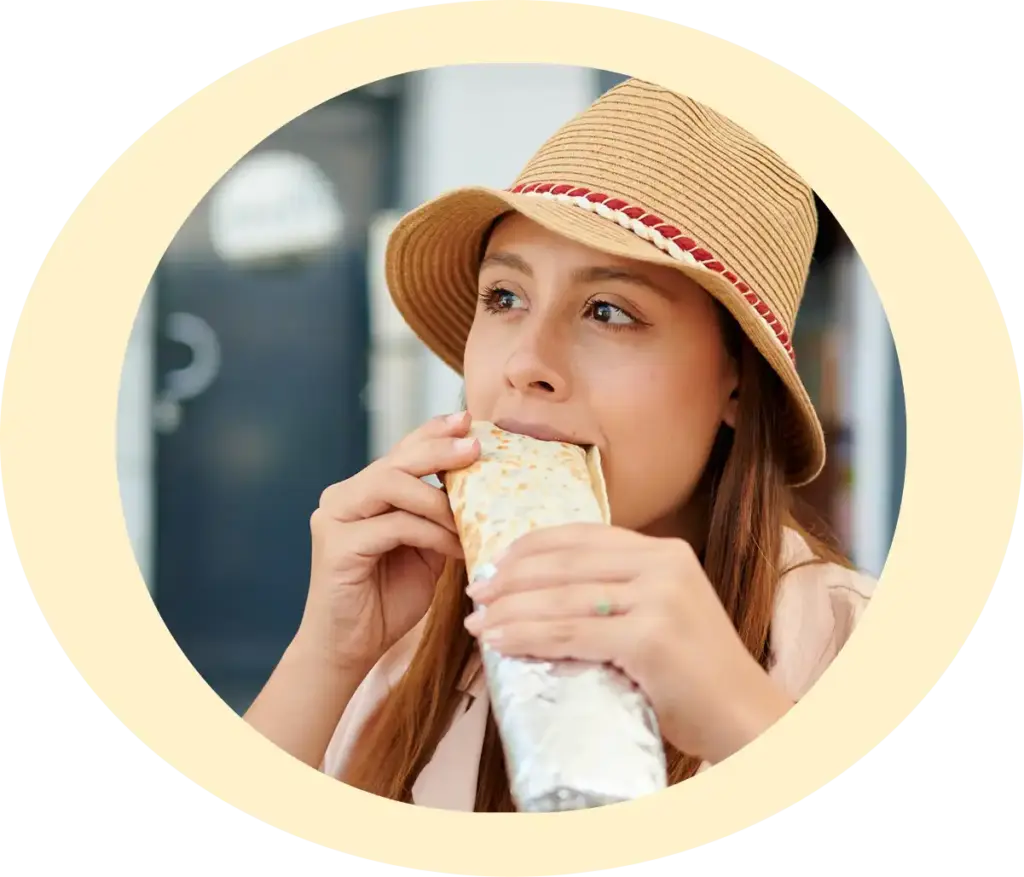 Kobieta w beżowym kapeluszu gryzie dużego kebaba, częściowo zawiniętego w folię, na tle otwartej kawiarni. Wygląda na zadowoloną i zrelaksowaną.